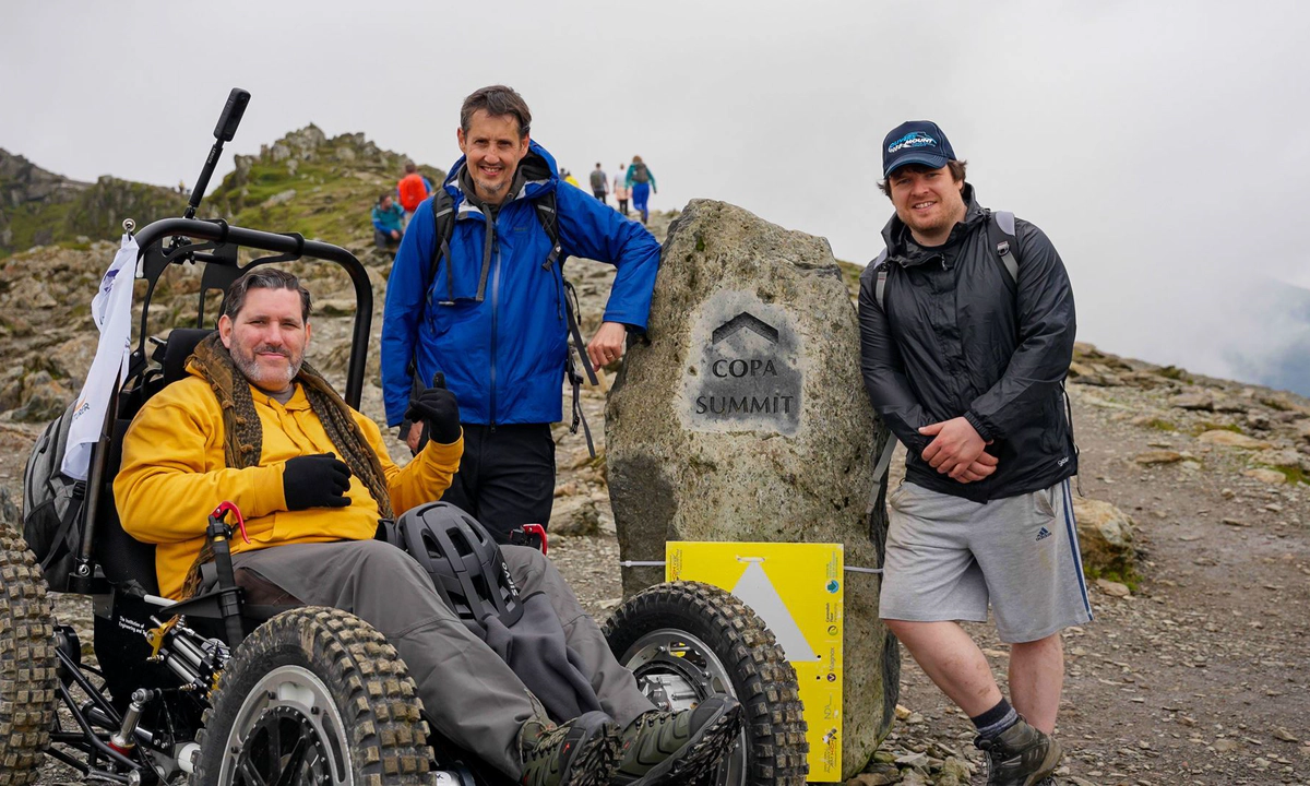 Nick Wilson in RockClimber beside the Copa Summit sign on Mount Snowdon. Nick is smiling, wearing a yellow hoodie, grey trousers, and black gloves.