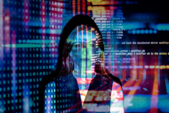 Photo of a woman standing in front of a wall facing the camera with colourful lights, lines of code, text and shapes projected onto her to convey tech.