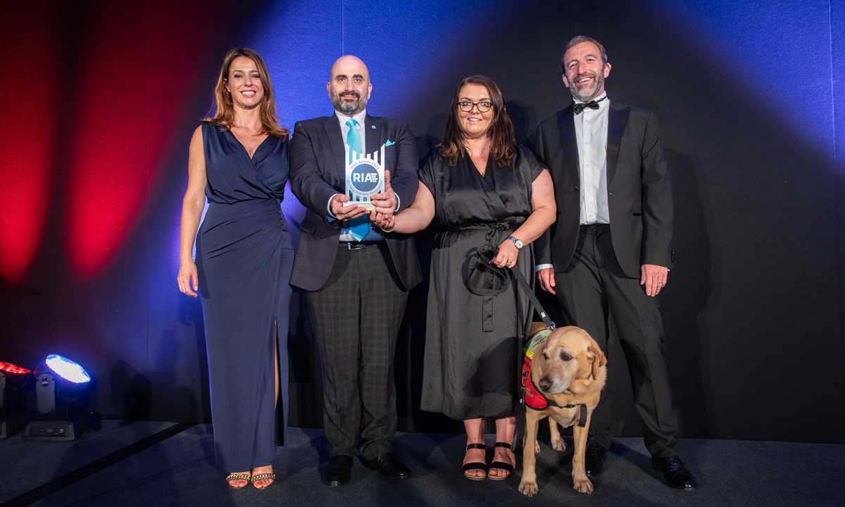 Transreport's Commercial Manager Joseph Mastracci, and Transreport's Accessibility and Inclusion Manager Emma Partlow holding the RIA RISE Award. Emma holds her assistance dog Luna on a black lead. Everyone in the photo is smiling proudly. 