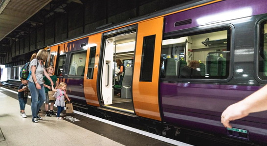 A purple and yellow West Midlands Railway train sat stationary at a platform at a train station, with a set of doors open and a group of adults and children about to board.