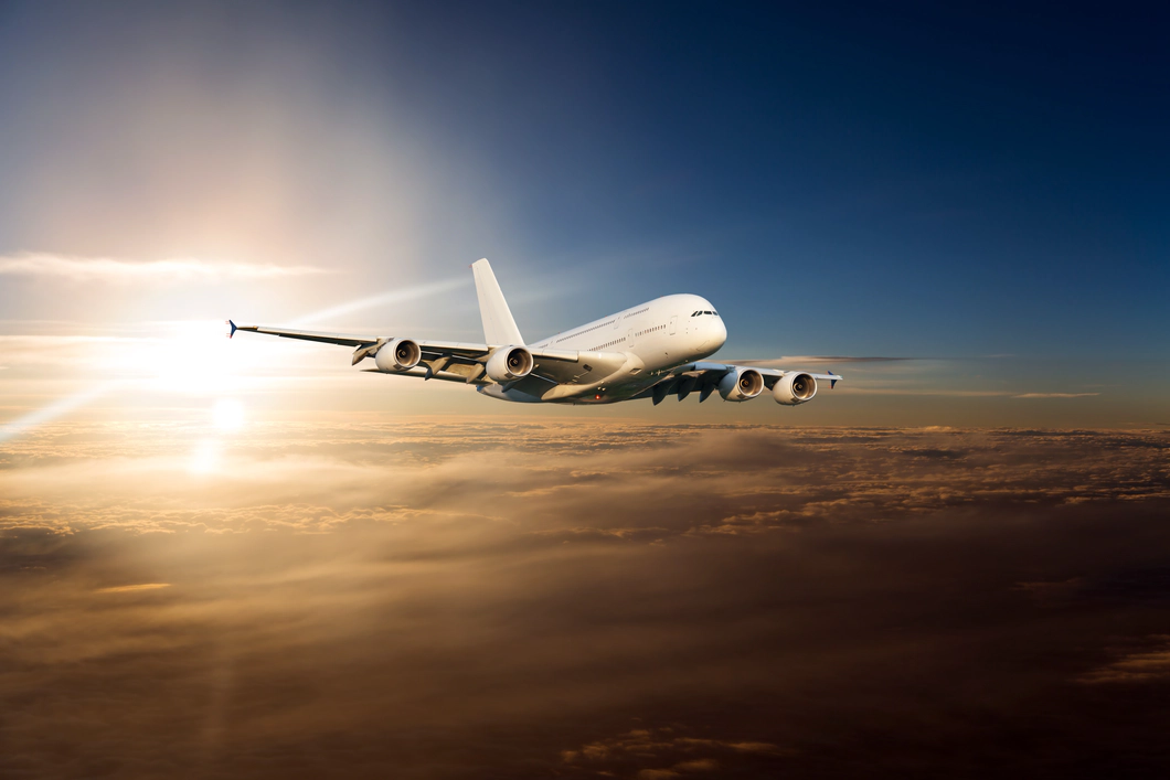 Photo of a white plane above a cloud bank being illuminated by a glowing sunrise below a dark blue sky.