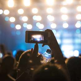 A photo of fans at a concert with one person taking a photo of the illuminated stage on their smart phone.