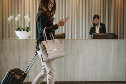 A woman smiling at her smart phone with a suitcase and handbag in front of a hotel check-in desk.