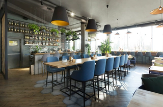 A photo of a restaurant with a long table, blue chairs, ceiling lamps, and wine bar.