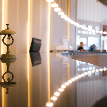Photo of a hotel check-in desk with a bell in the foreground and a customer speaking to a member of staff in the background.