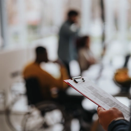 Photo with a blur effect of a restaurant. The person in focus holds a clipboard with information. In the background are three people including a man in a yellow jumper who is a wheelchair user.