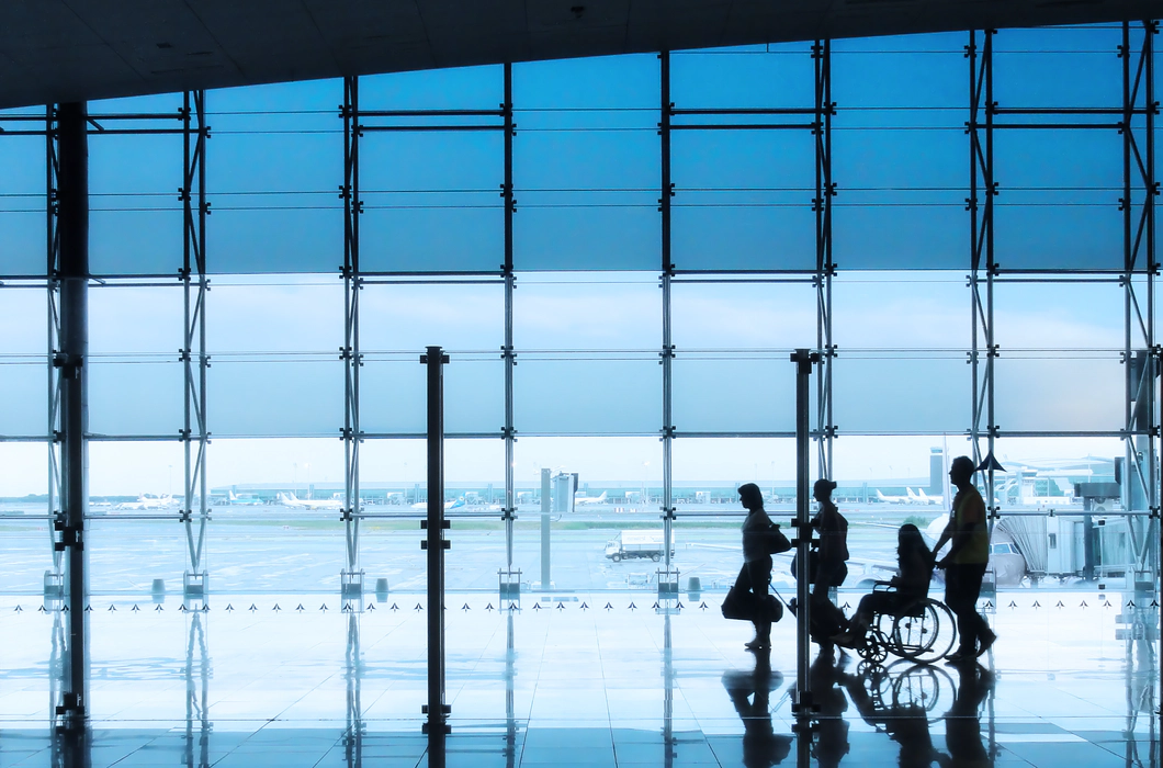 Four silhouettes of people in an airport setting, with a man and woman walking together with suitcases, followed by a wheelchair user being pushed by another person