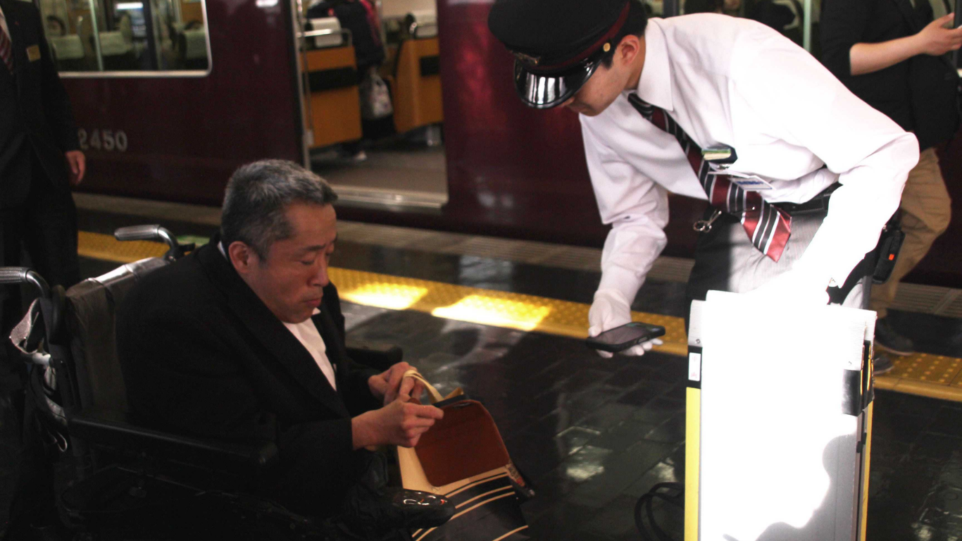 A photo of a man who is a wheelchair user being supported by a Hankyu staff member who is holding a smart phone and a wheelchair ramp.