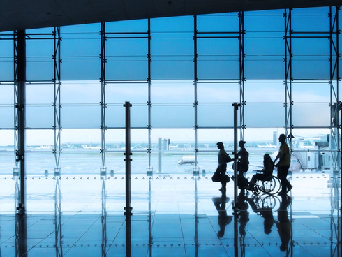 Four silhouettes of people in an airport setting, with a man and woman walking together with suitcases, followed by a wheelchair user being pushed by another person