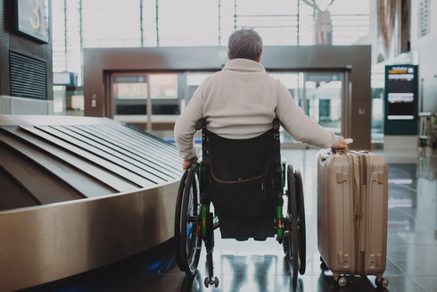 A man wearing a white jumper sitting in a wheelchair with his back to the camera, holding a suitcase on wheels in his right hand, in an airport.