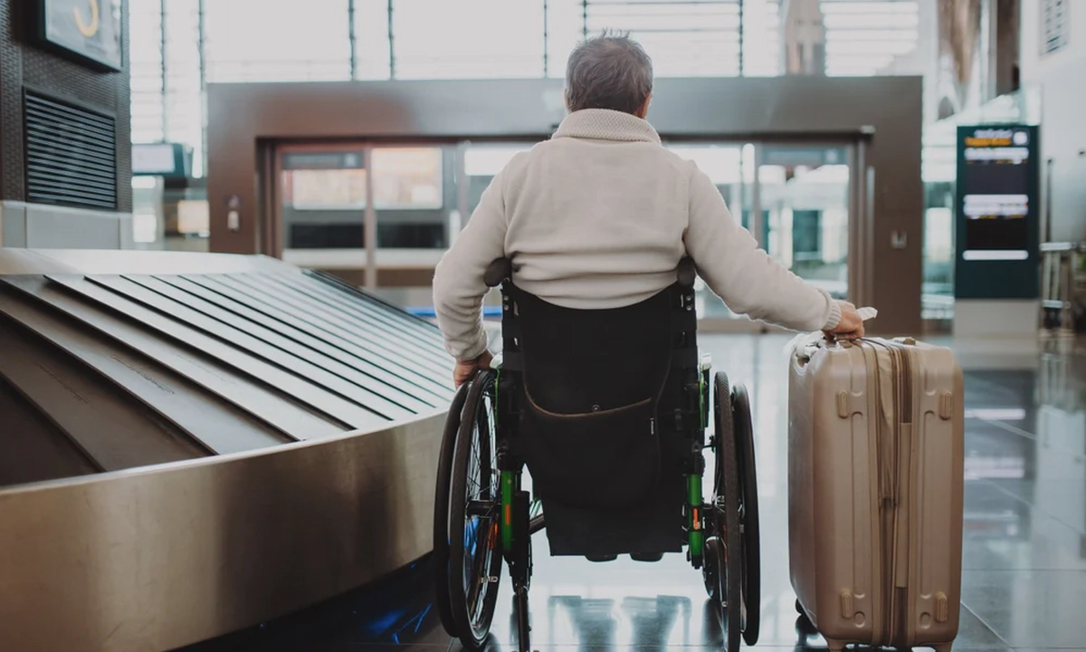 A man wearing a white jumper sitting in a wheelchair with his back to the camera, holding a suitcase on wheels in his right hand, in an airport.
