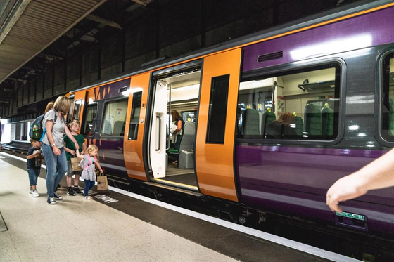 A purple and yellow West Midlands Railway train sat stationary at a platform at a train station, with a set of doors open and a group of adults and children about to board.